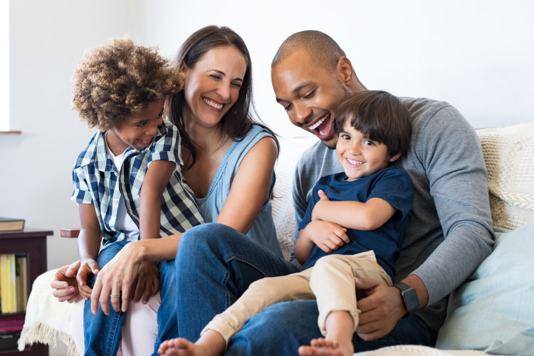 Multiracial family of a mom, dad, and two son smiling and laughing as they sit together on a couch