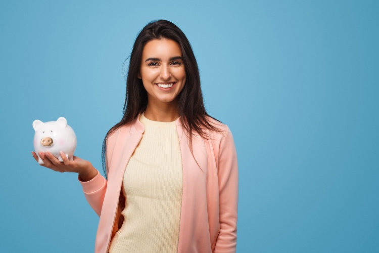 Brunette woman holds a piggy bank against a blue wall while wearing a pink cardigan