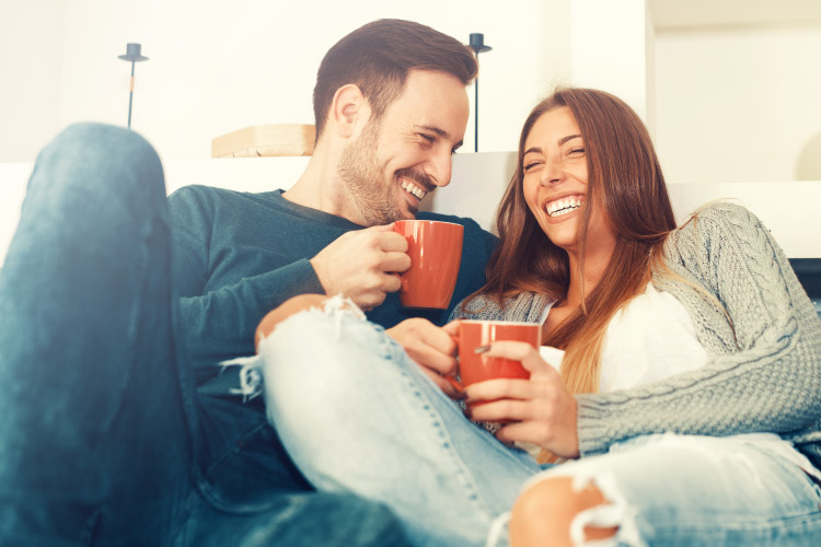 Brunette husband and wife drinking from mugs smile with their dental crowns while sitting on a couch