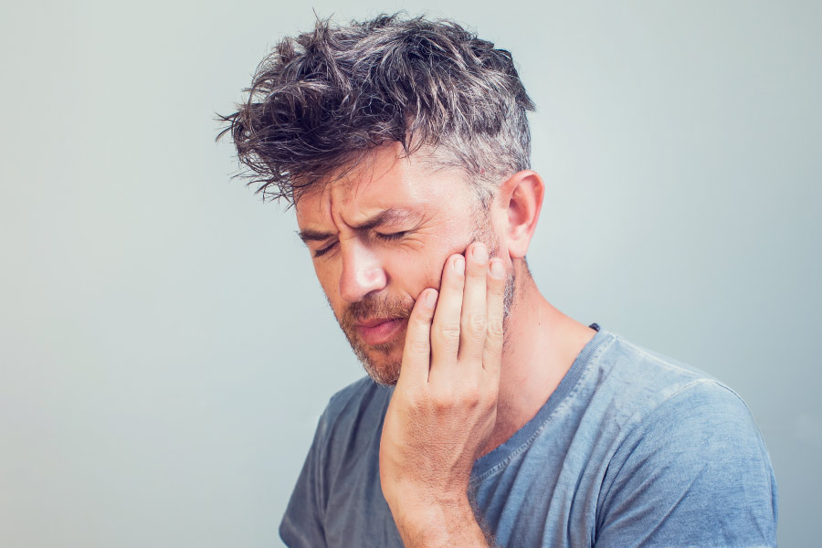 Middle-aged man in a gray t-shirt cringes in pain and touches his cheek due to a dental emergency