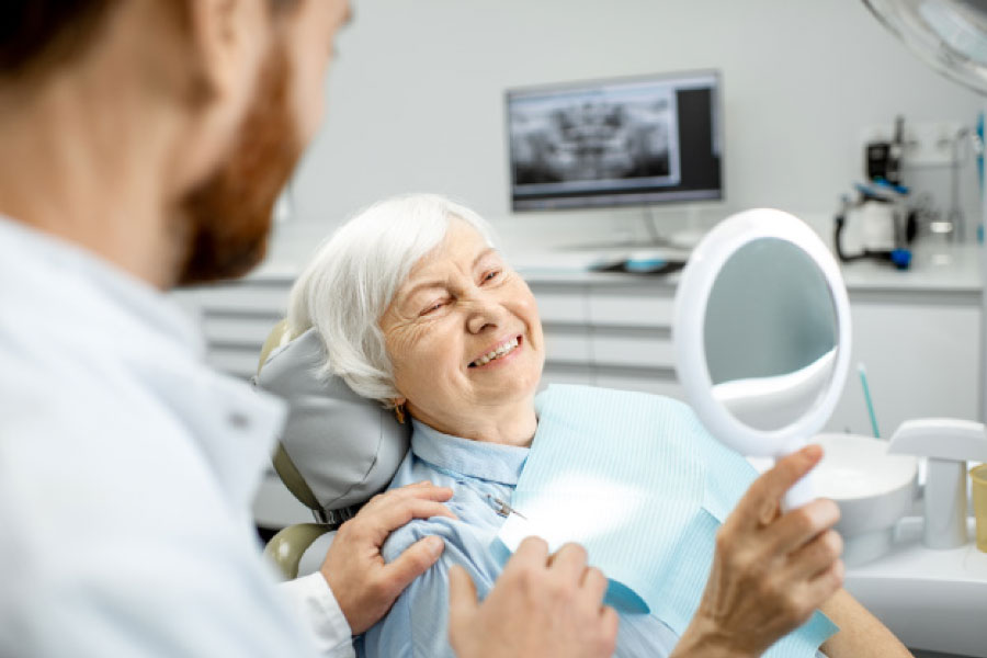 senior woman looks at her teeth in a handheld mirror as the dentist looks on